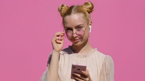 portrait of happy teen blonde girl in pink sunglasses touches hands with them smiling listens to music in airpods looks at camera on pink background slow motion in summer. emotions. positive mood