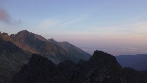 Wide-aerial-view-high-above-rocky-peaks-of-mountains-and-sunset-sky-background