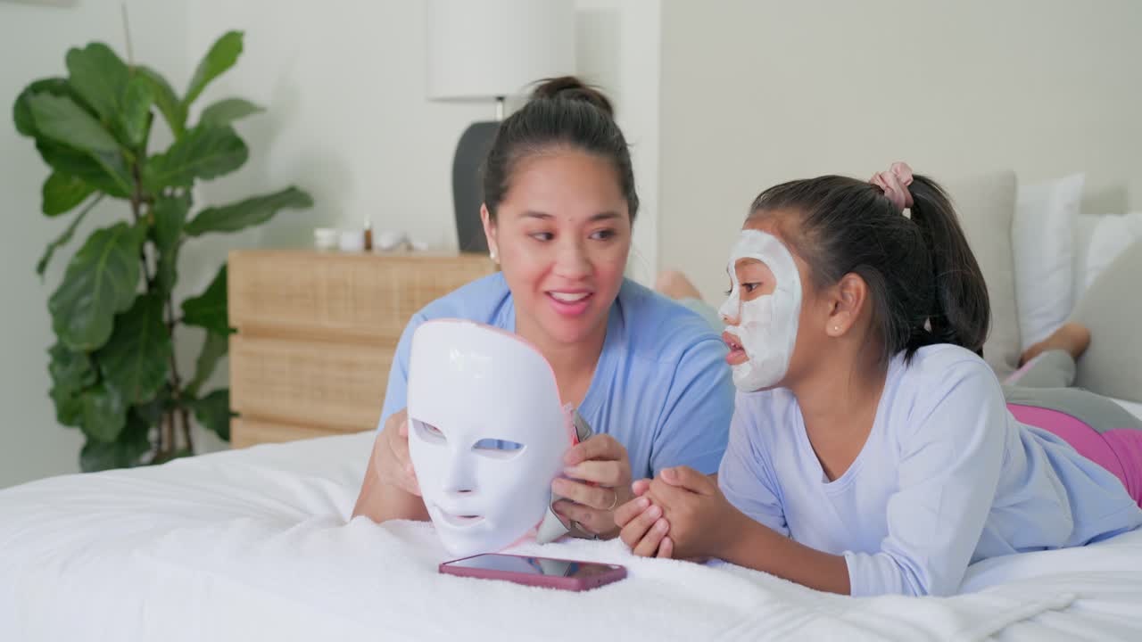 Asian mother demonstrating LED mask on bed with African American teen daughter wearing sheet mask