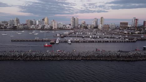 toma de seguimiento aéreo cinematográfico de lancha de pesca roja que ingresa al puerto de la ciudad de punta del este en uruguay con horizonte y rascacielos en segundo plano