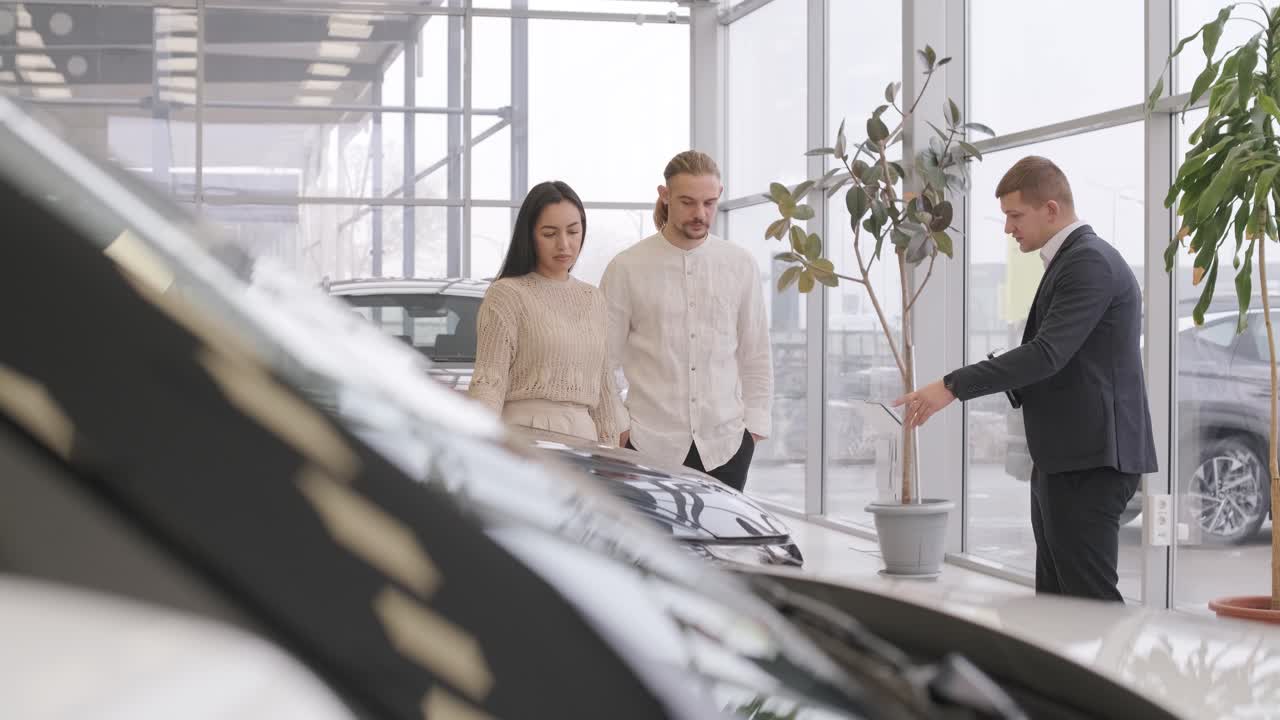 Young car salesman showing to young couple new automobile at dealership salon.