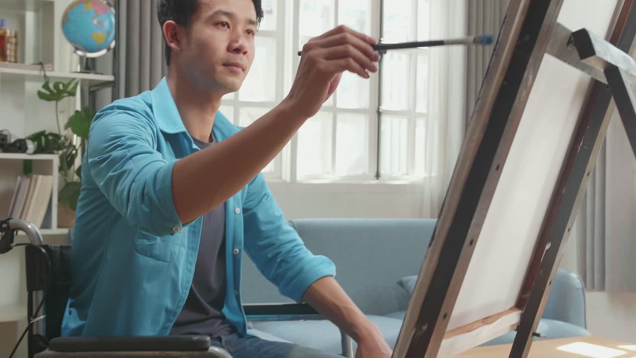 Asian Artist Man In Wheelchair Holding Paintbrush And Mixing It With Colour Before Painting On The Canvas