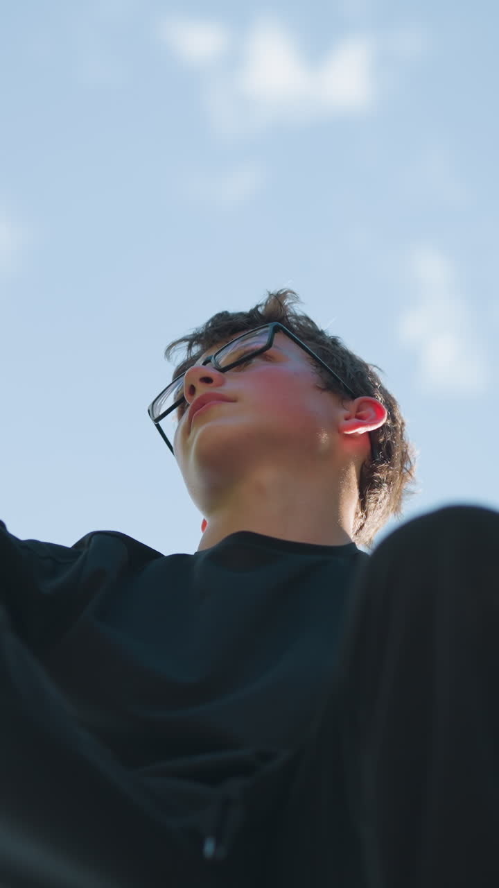 una vista hacia arriba de un niño sentado en un equipo de hierro azul al aire libre, sosteniendo la estructura con ambas manos mientras mira hacia la derecha