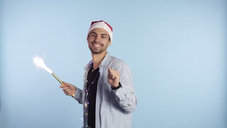 Positive-handsome-man-in-a-Christmas-red-hat-and-garland-on-neck-dances-and-waving-hand-with-a-burning-cracker,-celebrating-Christmas-or-New-Year.-Background-isolated-on-blue