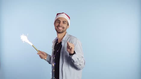 positive handsome man in a christmas red hat and garland on neck dances and waving hand with a burning cracker, celebrating christmas or new year. background isolated on blue