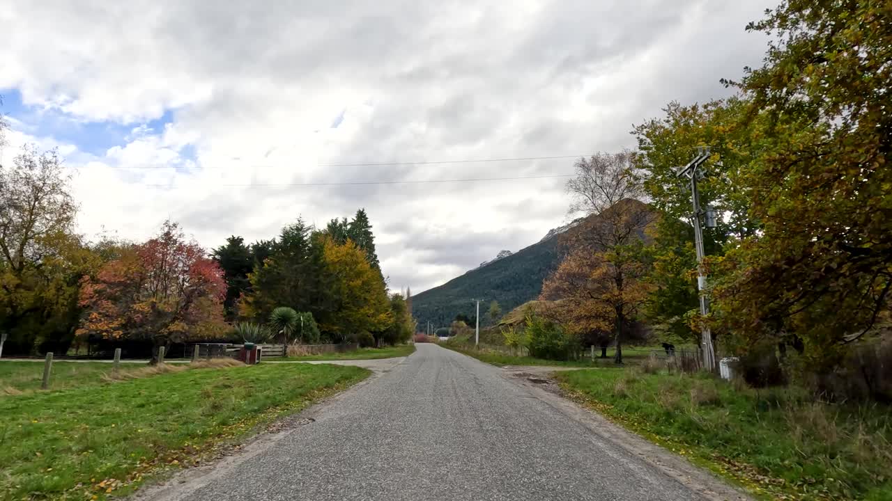 Forward-moving camera captures autumn trees, gravel road, and mountain landscape under overcast daylight