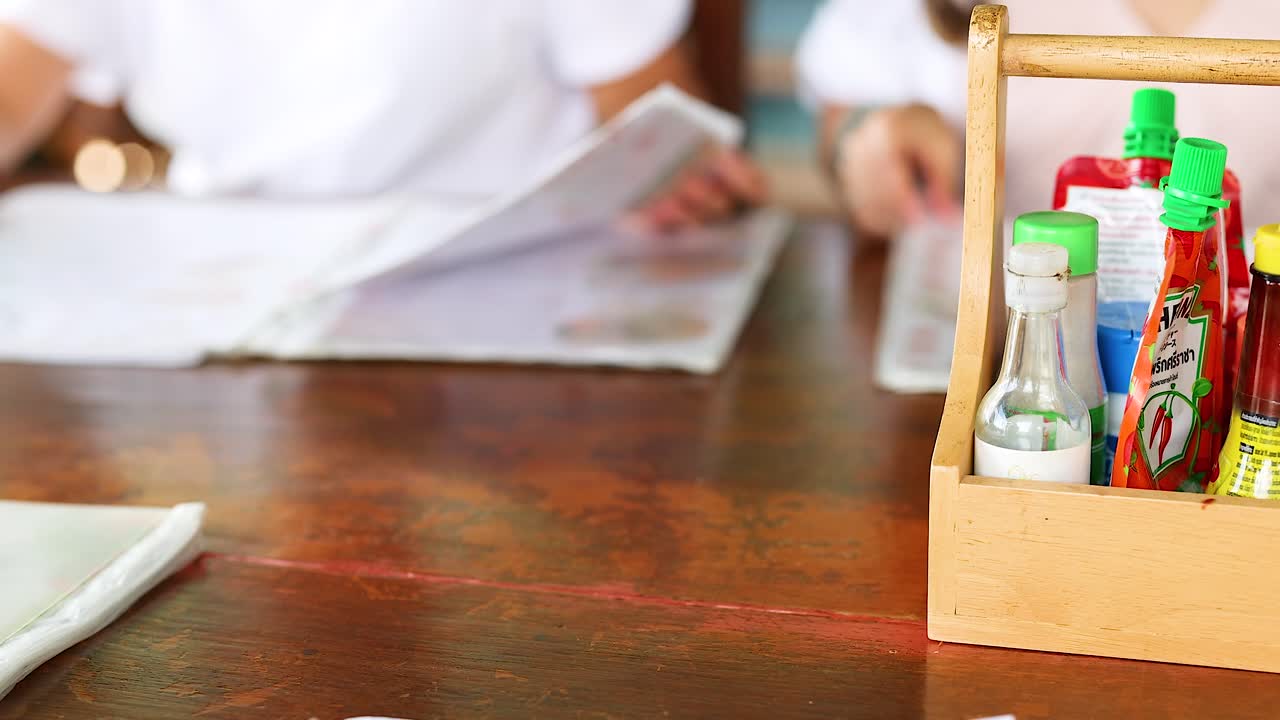 A person reviews a menu at a Phuket restaurant table with various condiments in focus. Bright, natural lighting