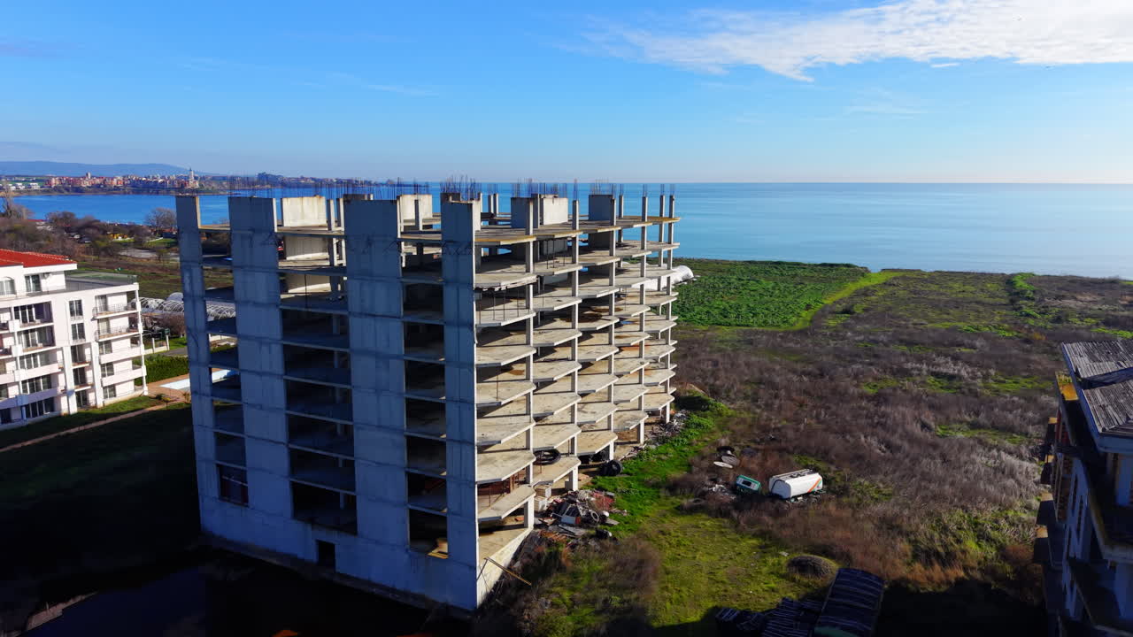 Vista posteriore dello scheletro di un alto edificio in cemento incompiuto con vista sul mare e su un paesaggio con serre. Aerea