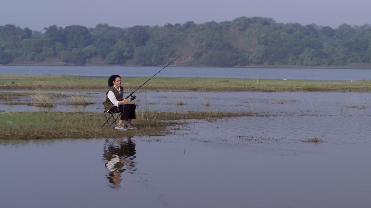 Seorang wanita Asia Selatan memancing di tepi danau yang tenang, duduk di kursi dan memegang pancing, menikmati alam pedesaan yang damai di luar ruangan dalam pemandangan siang hari.