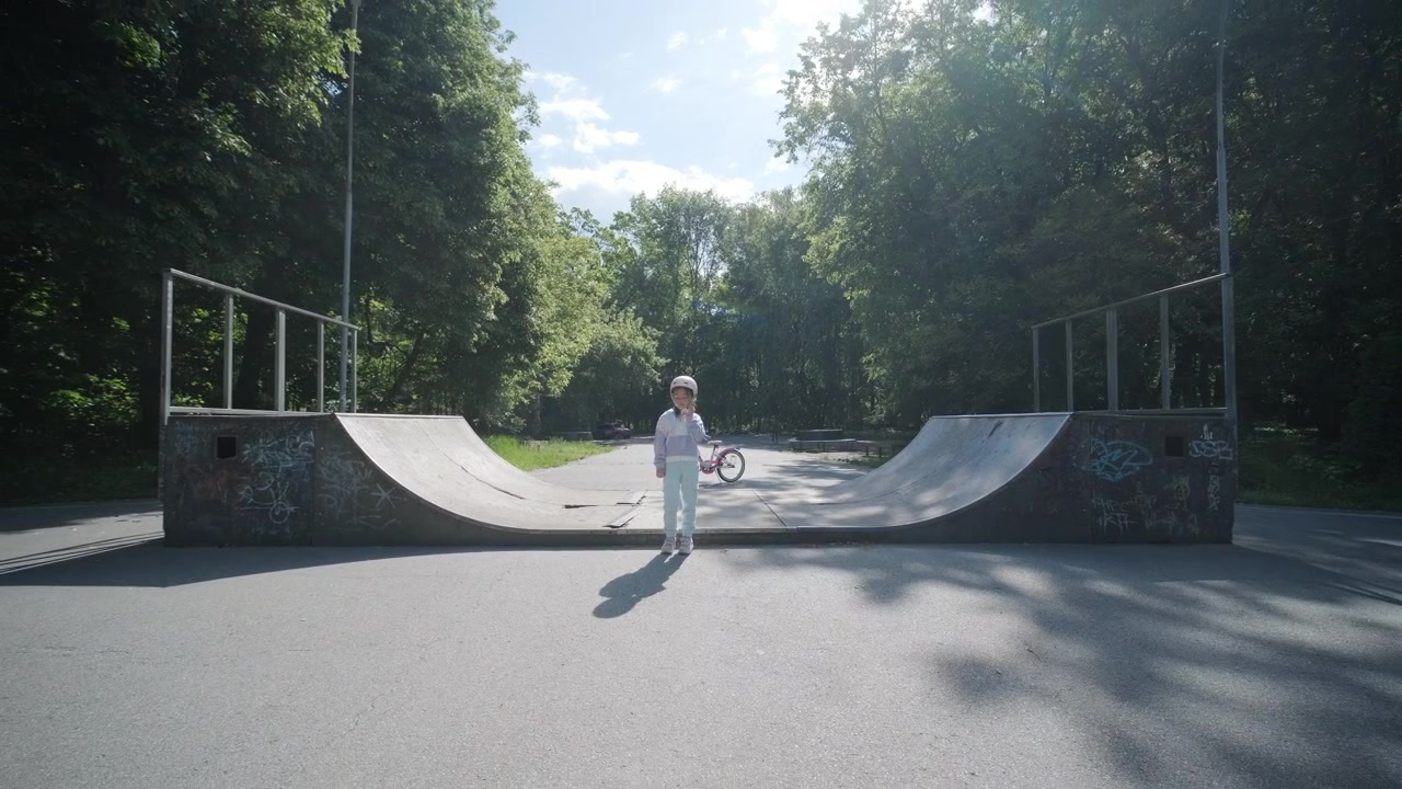 Giovane ragazza con casco in piedi in uno skate park soleggiato con half-pipe