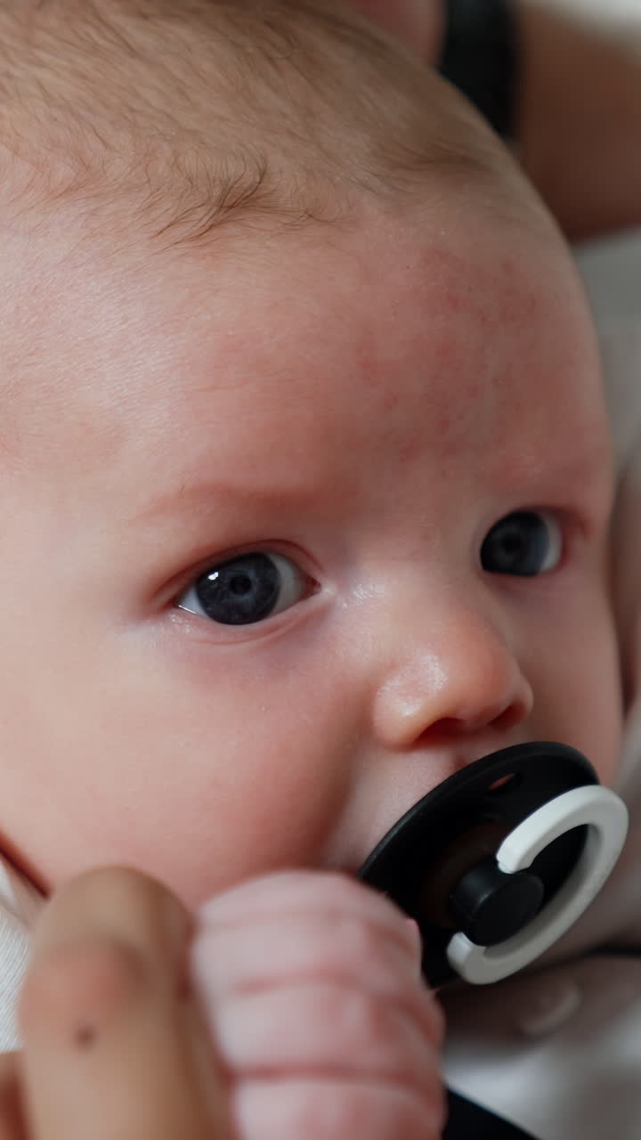 Beautiful grey-eyed Caucasian newborn in mom's arms. Baby lies peacefully suckling the pacifier. Close up. Vertical video