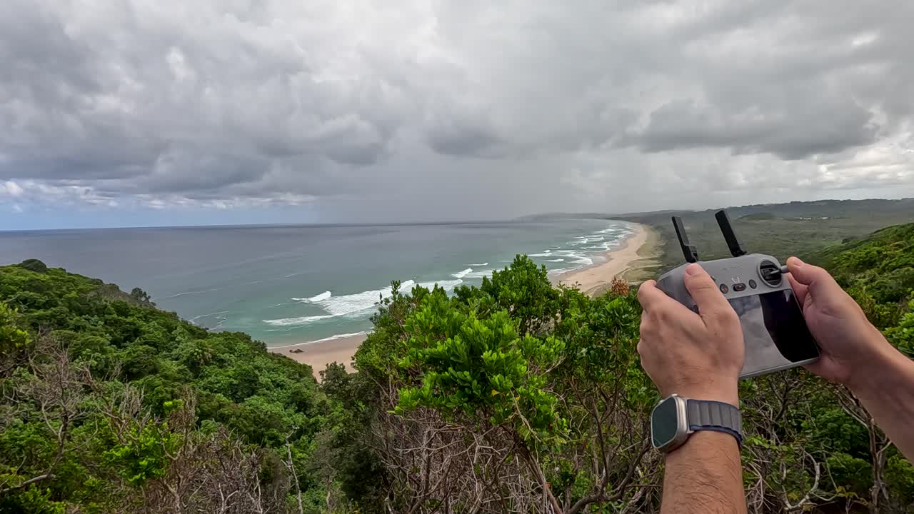A person operates a drone over lush greenery and ocean views under cloudy skies, capturing scenic coastal beauty