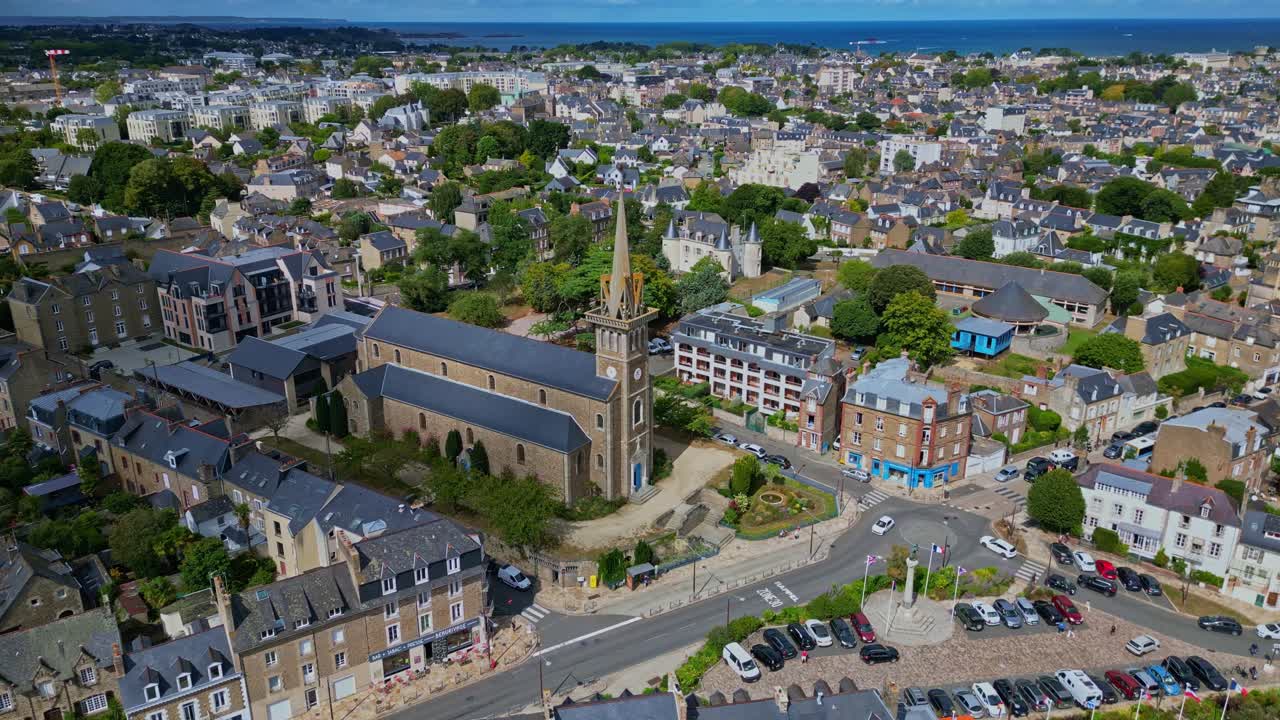 An orbiting drone shot shows Notre-Dame d’Émeraude Church in Dinard, surrounded by city buildings, streets, and traffic, with the coastline visible in the distance