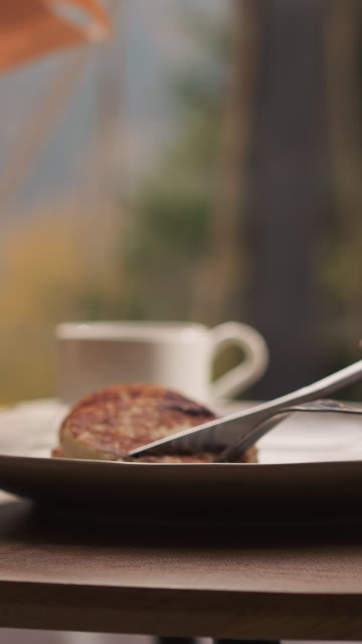 Lady uses knife and fork at breakfast in cottage. Woman has pancakes for morning meal by panoramic window with view on nature. Woman cuts pancakes