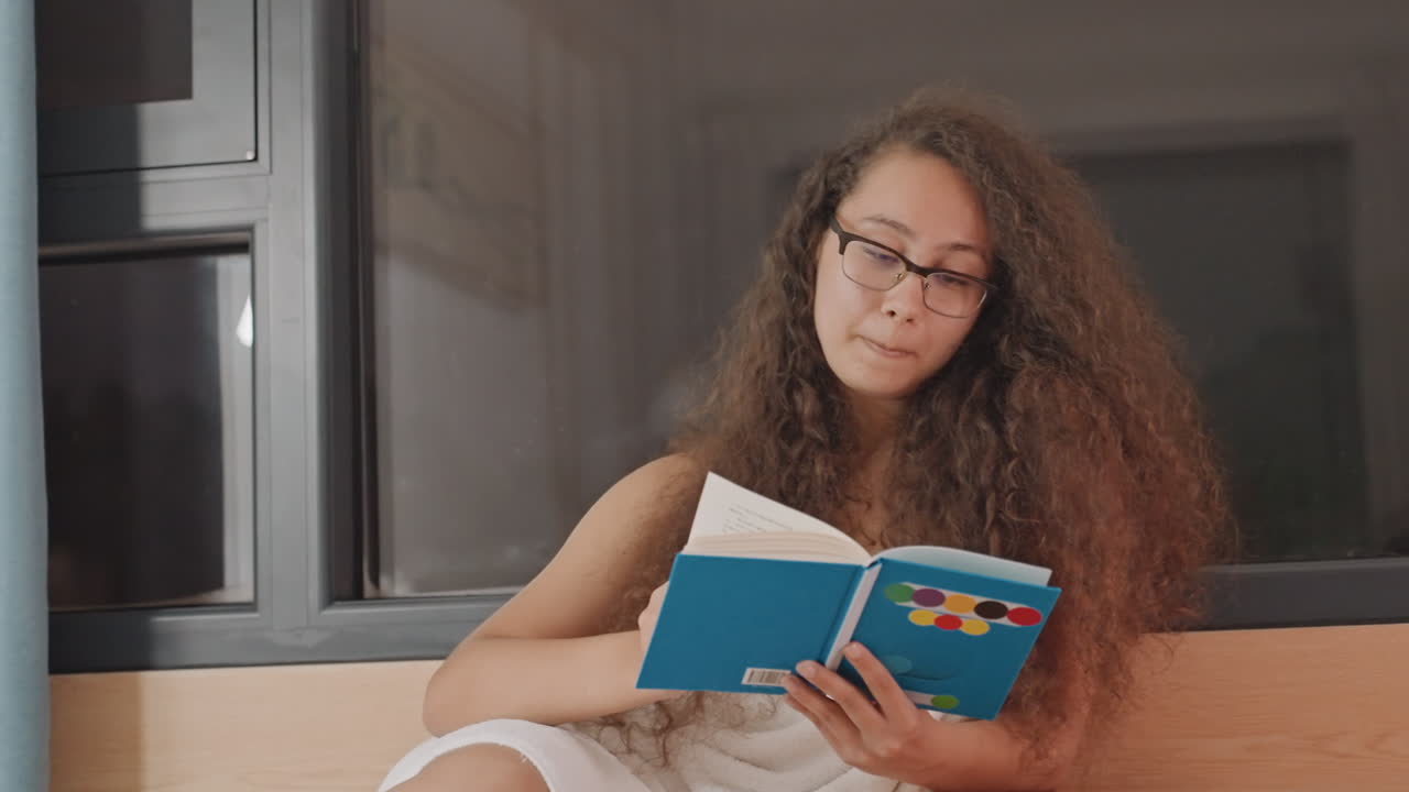 Une femme lisant dans le renfoncement d'une fenêtre d'hôtel, tournant les pages d'un livre bleu avec intention. Un banc en bois chaud et un fond de fenêtre sombre, suggérant une solitude nocturne et une réflexion créative. Des gestes doux suggèrent une écrivaine.