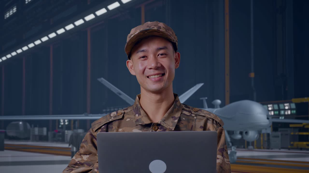 Pria Prajurit Asia Menggunakan Laptop dan Tersenyum Sambil Berdiri dengan Drone Militer Modern di Hangar Pemeliharaan.