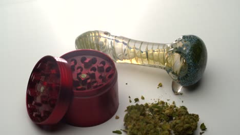 closeup view of a glass pipe on a white table as the camera pulls back to reveal an open red grinder and marijuana buds