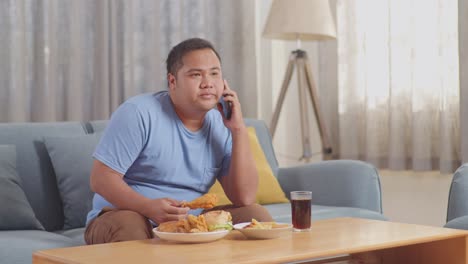 a fat asian man talking on smartphone while eating fast food having fried chicken on a sofa in the living room at home