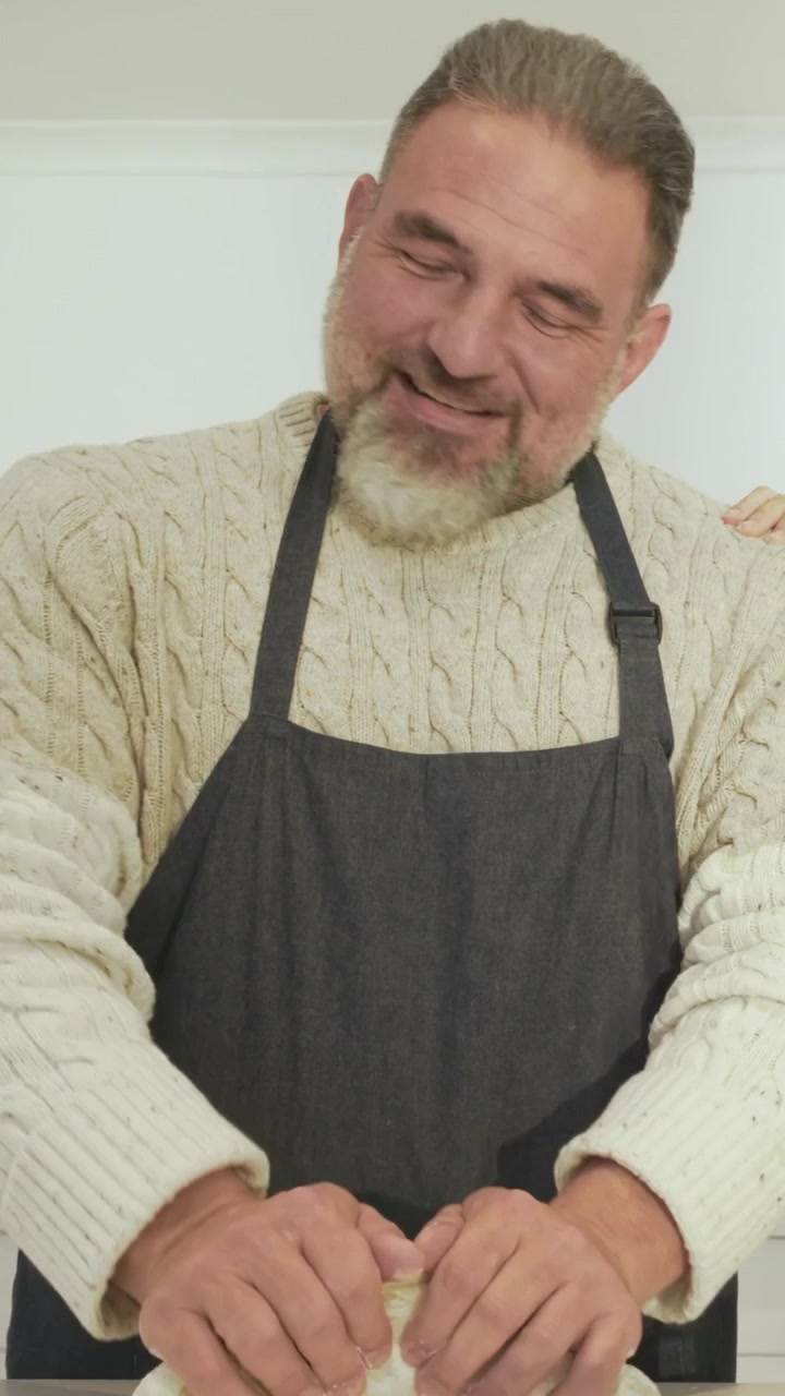 Vertikales Video: Mann knetet Teig in der Küche in Schürze, spürt die Hand des Partners auf der Schulter und lächelt.