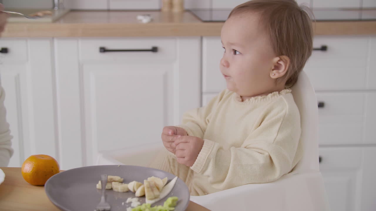Cute Little Girl Sitting In High Chair In The Kitchen While Her Unrecognizable Mom Feeding Her