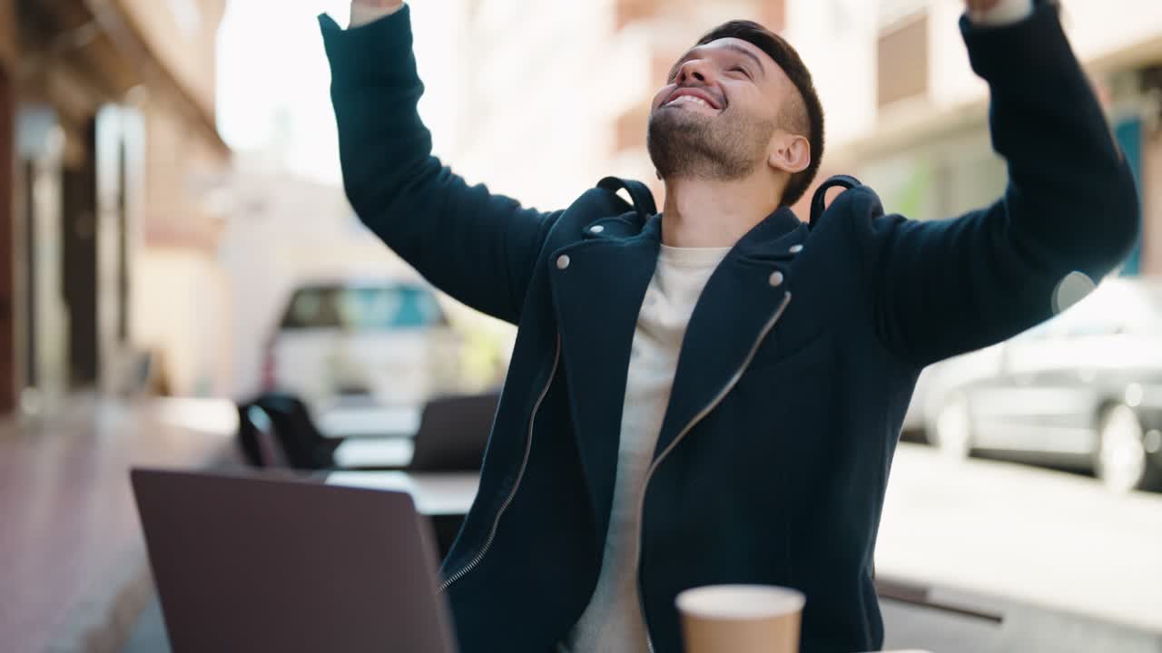 joven hispano con expresión de ganador usando una computadora portátil en la terraza de la cafetería