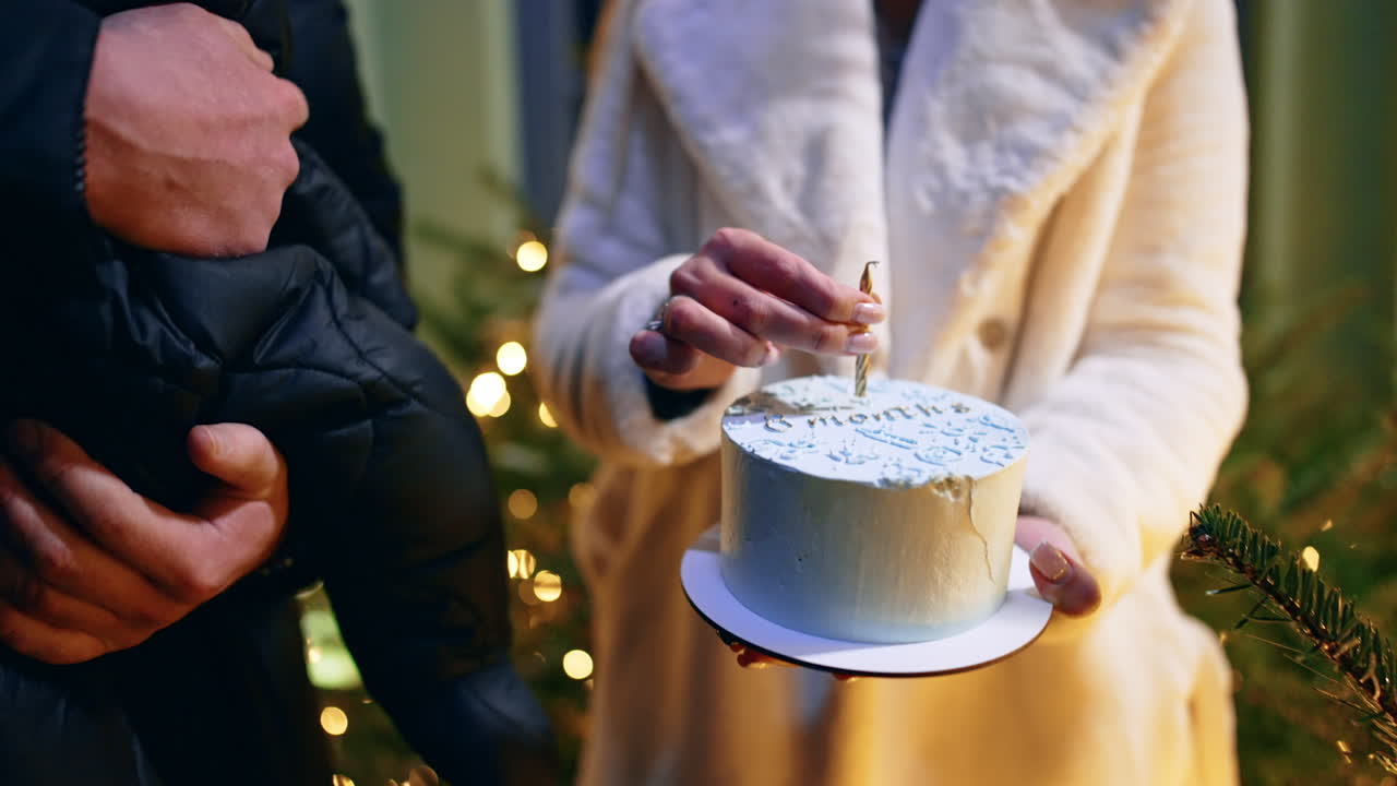 Unrecognized woman in white coat holds a white cake and a candle over it. Man holding a baby stands beside. Close up.