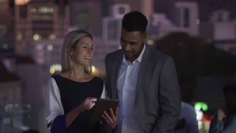 video of two happy diverse male and female colleagues talking and using tablet at night in office