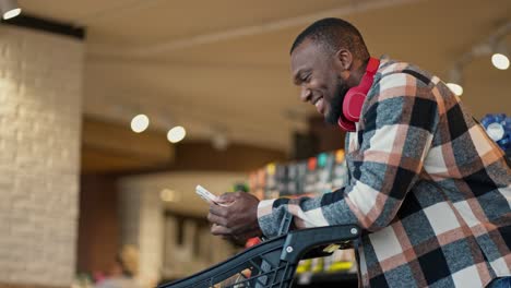 a happy and joyful man with black skin and a short haircut and beard in a plaid shirt and wireless headphones looks at the list of products on his phone that he needs to buy while going to the supermarket with a cart