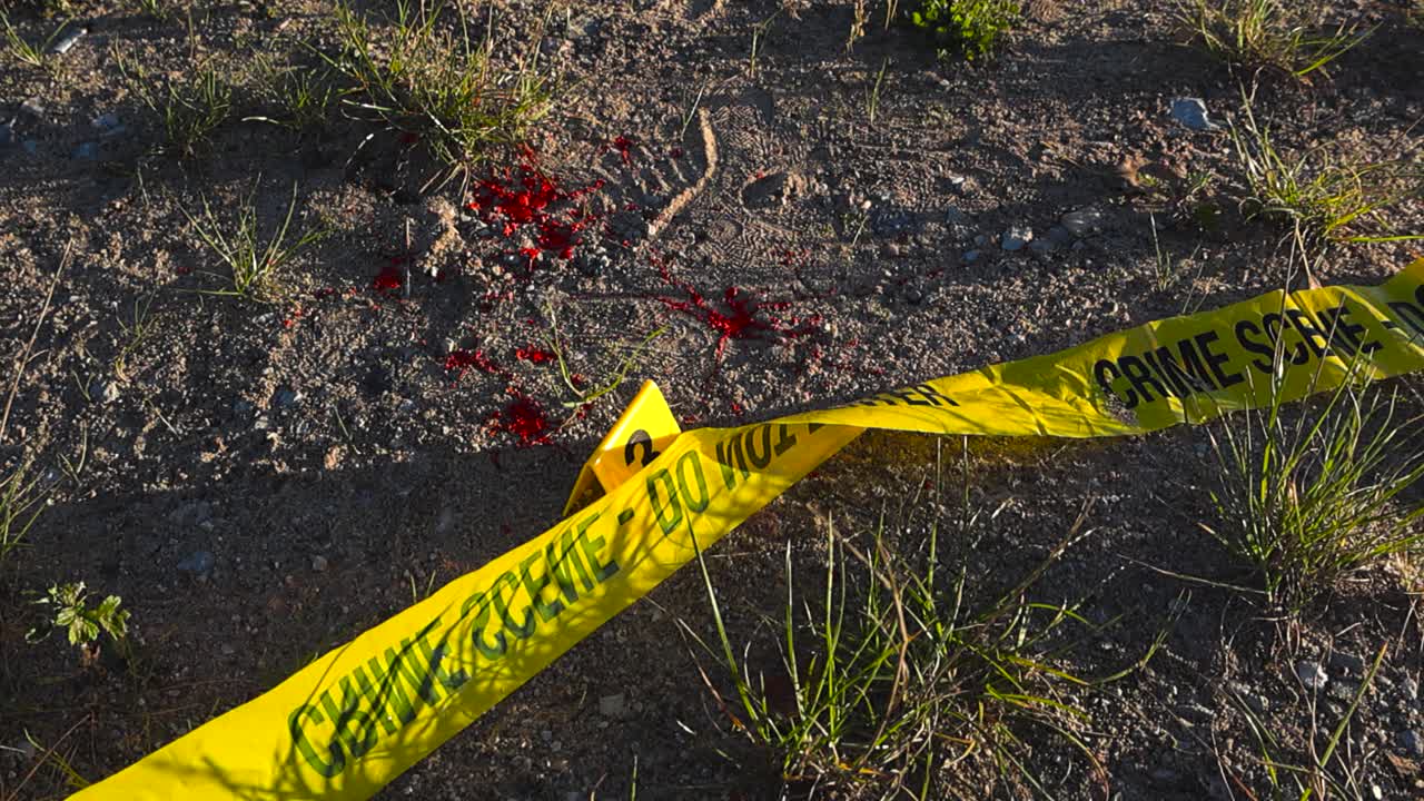 Top down view of red blood spatter on dirt ground with a yellow police Crime marker and forensic investigation yellow crime scene tape on the ground next to it, appers to be a cold case, abandoned