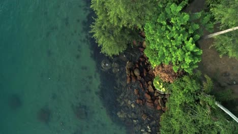 drone flight clip from above of a stony natural coastline and a small house at the beach with a wooden landing stage, two boats and few people