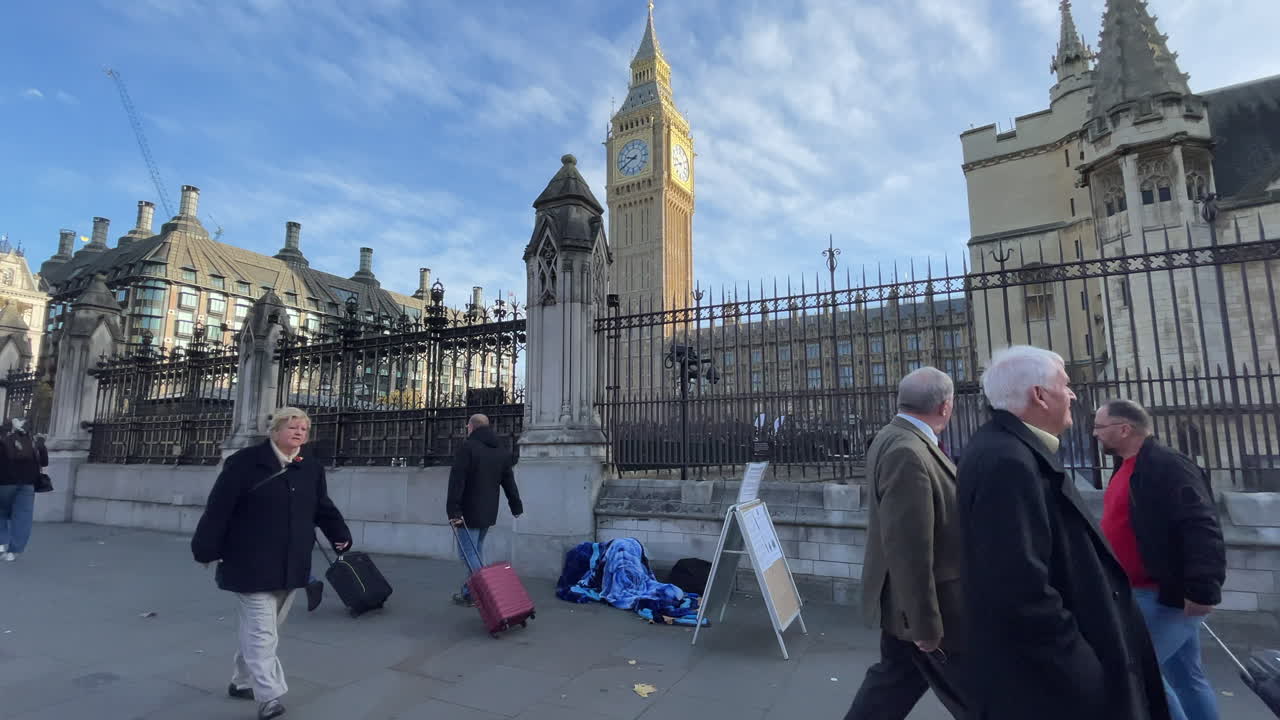 People walk past a sleeping homeless person underneath a blue blanket in front of the Houses of Parliament and the Elizabeth Tower that is home to the Big Ben bell on a clear winter morning.