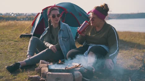 happy girls drink tea at burning bonfire near blue tent