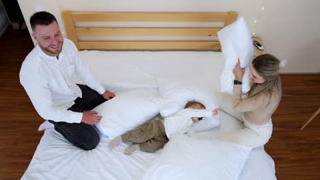 adorable caucasian toddler with pacifier lies relaxed in the pillows. mom and dad kick the son gently and playfully with pillows. top view.