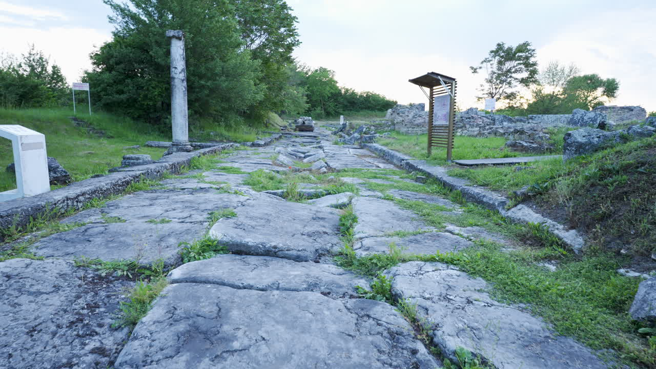 Slow motion push in shot along a Roman stone road with grass between the stones at Nicopolis ad Istrum, surrounded by greenery and cloudy sky
