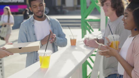 blonde girl bringing pizza to share with her friends while standing at an outdoor table in the street, drinking cold drinks and chatting together