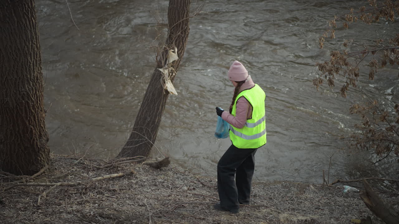 Woman in bright reflective vest picks up trash along riverbank during environmental cleanup, holding blue plastic bag, wearing pink beanie and gloves, helping remove litter from muddy shoreline near flowing water