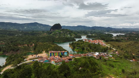 Toma-Aérea-De-Seguimiento-Frente-A-Un-Pueblo-Con-El-Fondo-De-La-Roca-De-Guatape-En-Colombia.