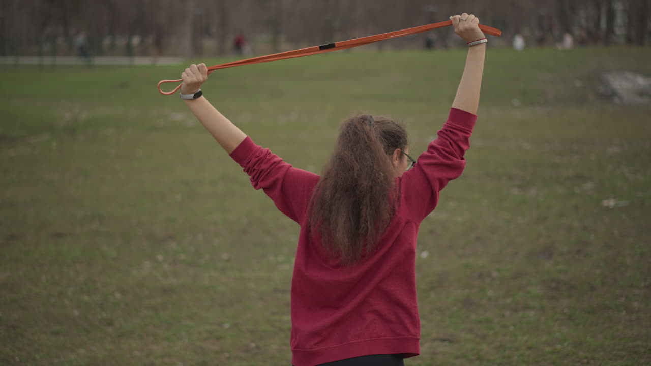Asian Woman Stretches, Open Park Features Woman Performing Stretches, Back View Of Woman Doing Band Stretches In Park, Female Asian Individual Engaging In Mobility And Posture Exercises Outdoors