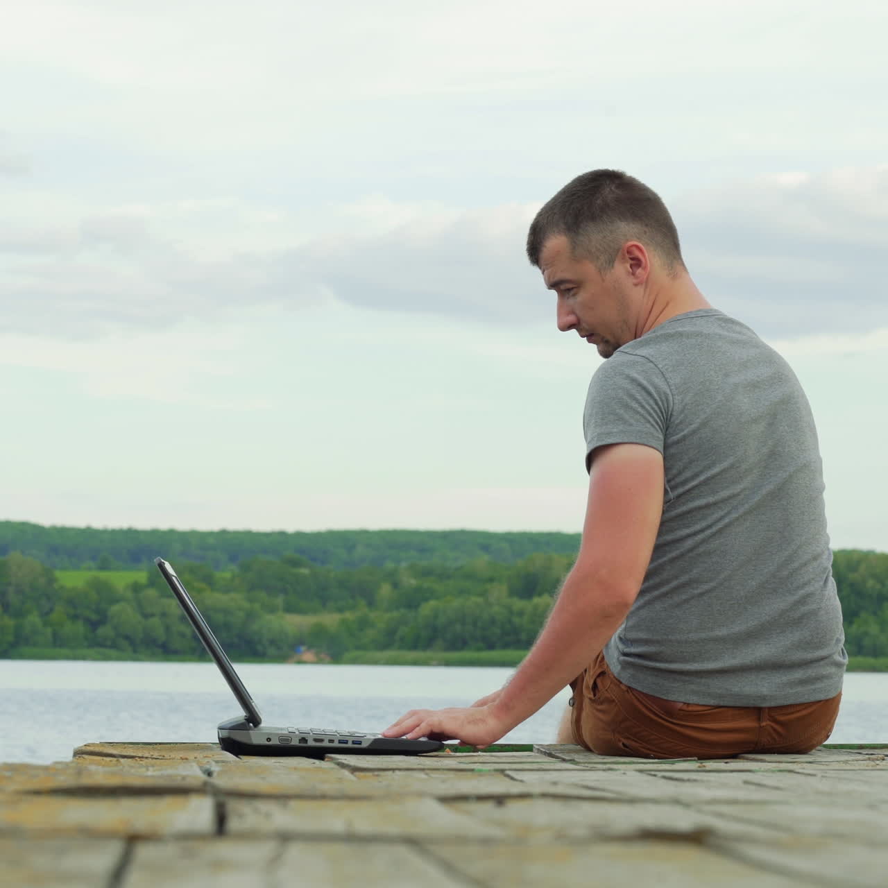 Young male sitting at the edge of the bridge and opens a laptop and started working near the river. Man in a grey t-shirt working on nature with modern gadget