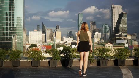 una mujer joven en un hotel en la azotea camina lentamente hacia el impresionante horizonte de bangkok, tailandia