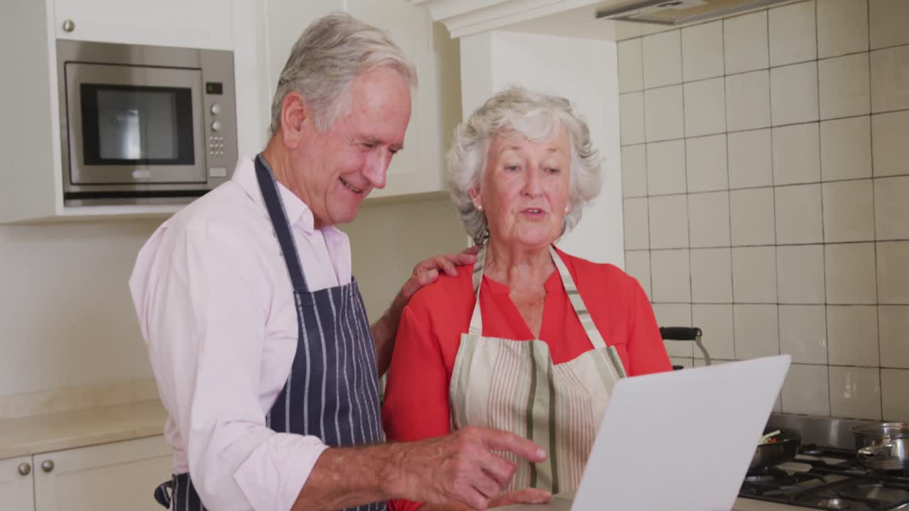 feliz pareja de adultos mayores caucásicos en la cocina usando delantales usando computadora portátil antes de preparar la comida