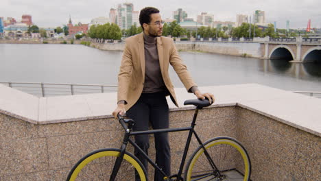 smiling american man looking at the camera and holding his bike while leaning against wall in the city