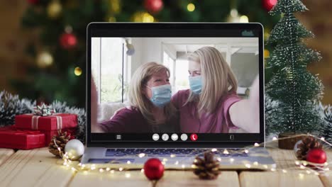 Madre-E-Hija-Con-Máscaras-En-Videollamada-En-Computadora-Portátil,-Con-Adornos-Navideños-Y-árbol