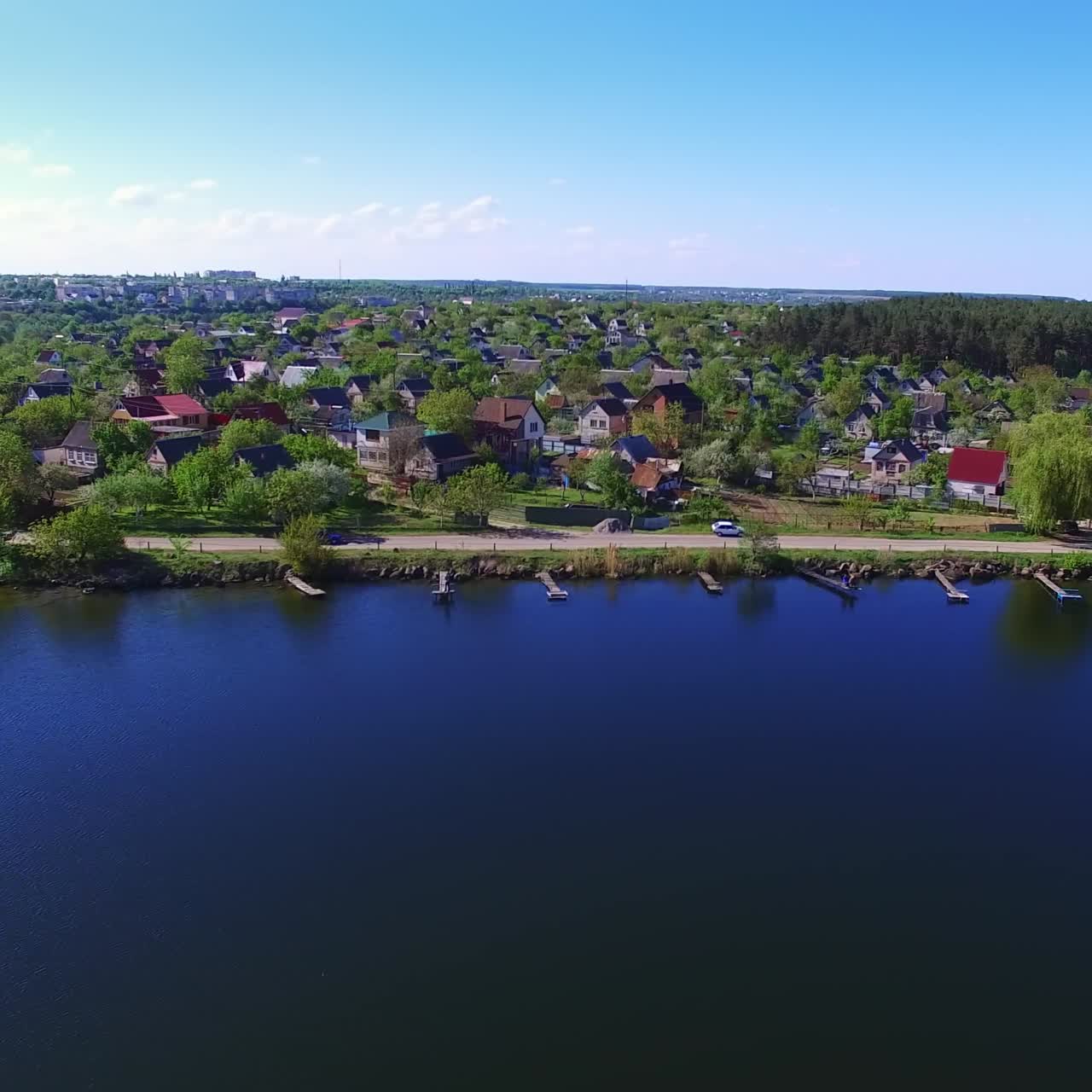 Green rural area locating on the river bank. Drone footage over the water along the waterfront with car road. Sunny daytime backdrop