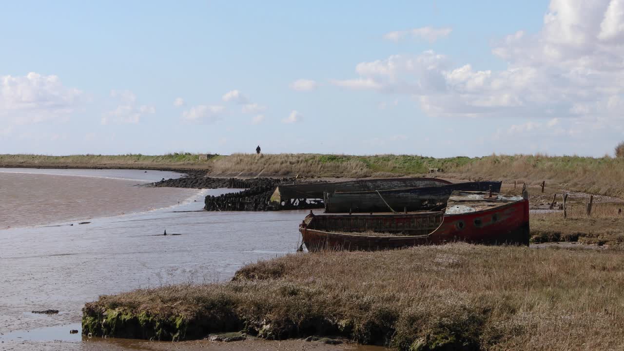 Relitto di barca sulla riva a Orford, sulla costa del Suffolk, Regno Unito, con una figura che si allontana in lontananza.