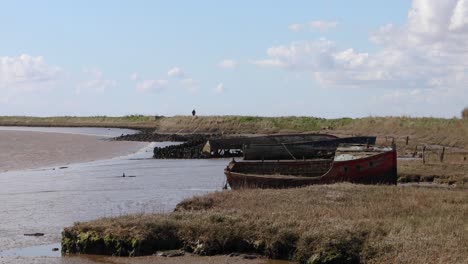 boat wreck on the shore at orford on the suffolk coastline, united kingdom, with figure walking away in the distance