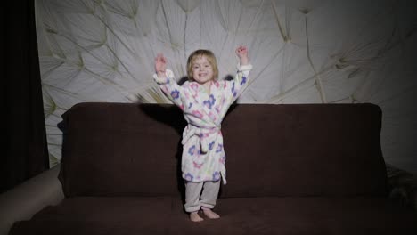 Little-cute-girl-with-blond-hair-jumps-on-sofa.-Bathrobe-clothes.-Happy