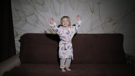 little cute girl with blond hair jumps on sofa. bathrobe clothes. happy