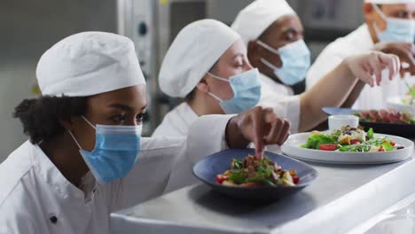 Diverse-group-of-chefs-wearing-face-masks-garnishing-dishes-in-restaurant-kitchen