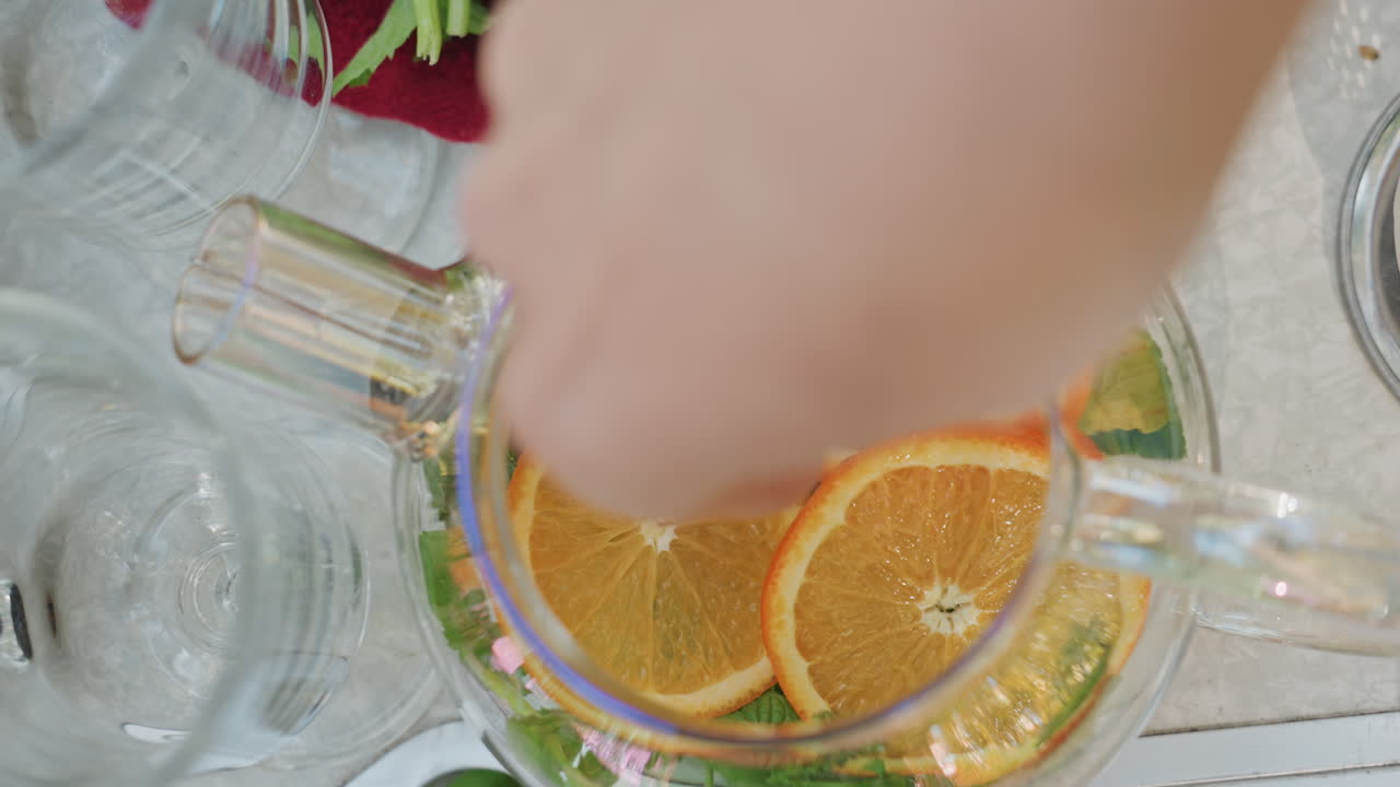 Mano colocando rodajas de naranja en una tetera, hierbas y tetera de cristal en una mesa al aire libre, ambiente de suave crepúsculo, vapor cálido y atmósfera resplandeciente de campamento, relajación comunitaria y bebida relajante para la noche.